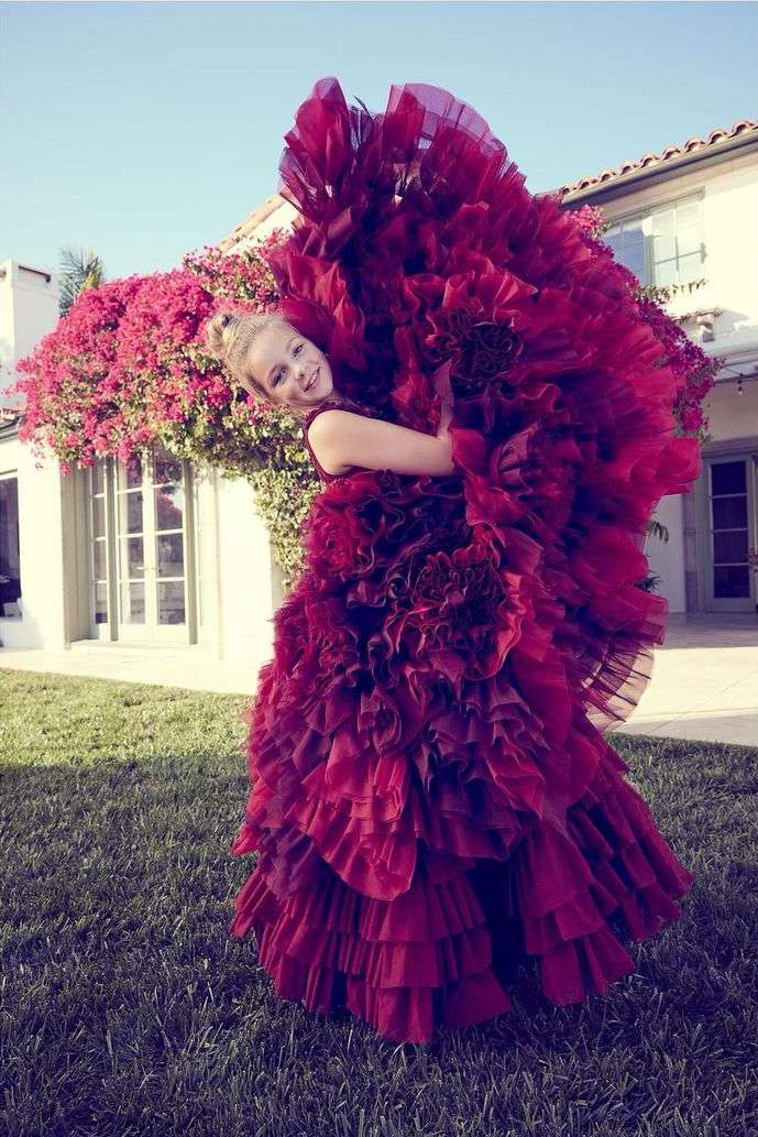 A blonde child stands on a lawn in a very large, red ruffled dress with a red flower bush and a white home in the background.