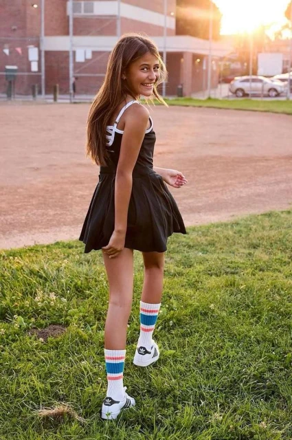 Young girl in black dress and striped socks poses outdoors with a smile, looking back over her shoulder at the camera during golden hour.