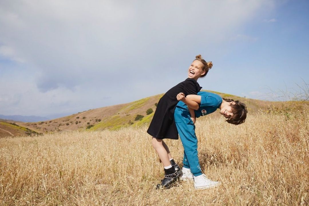 A laughing girl and boy, wearing Champion brand clothing, are outdoors in a field with rolling hills and a blue sky with clouds.