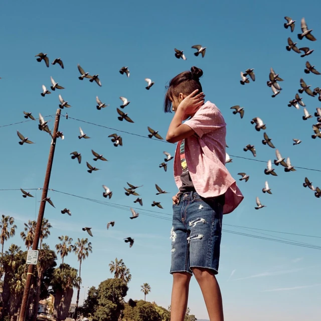 Young person surrounded by flock of pigeons in an urban setting on a sunny day, wearing denim shorts and a pink shirt against a bright blue sky.