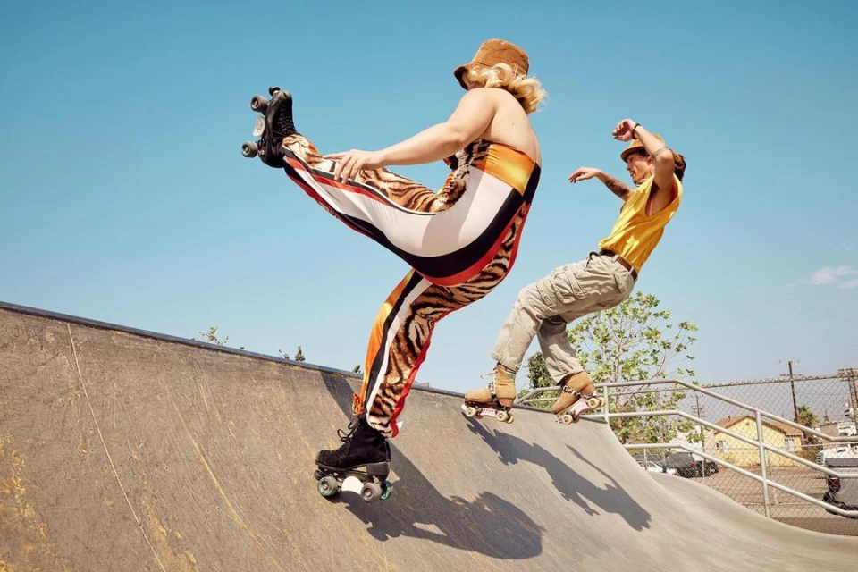 Two people roller skating at an outdoor skate park, one performing a stunt kick, with a blue sky and concrete ramps.