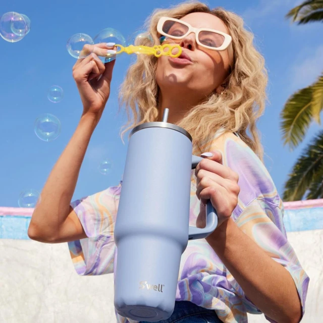 Blonde woman outdoors blowing bubbles near a blue cup with a straw, wearing sunglasses and a patterned top, with blue sky.