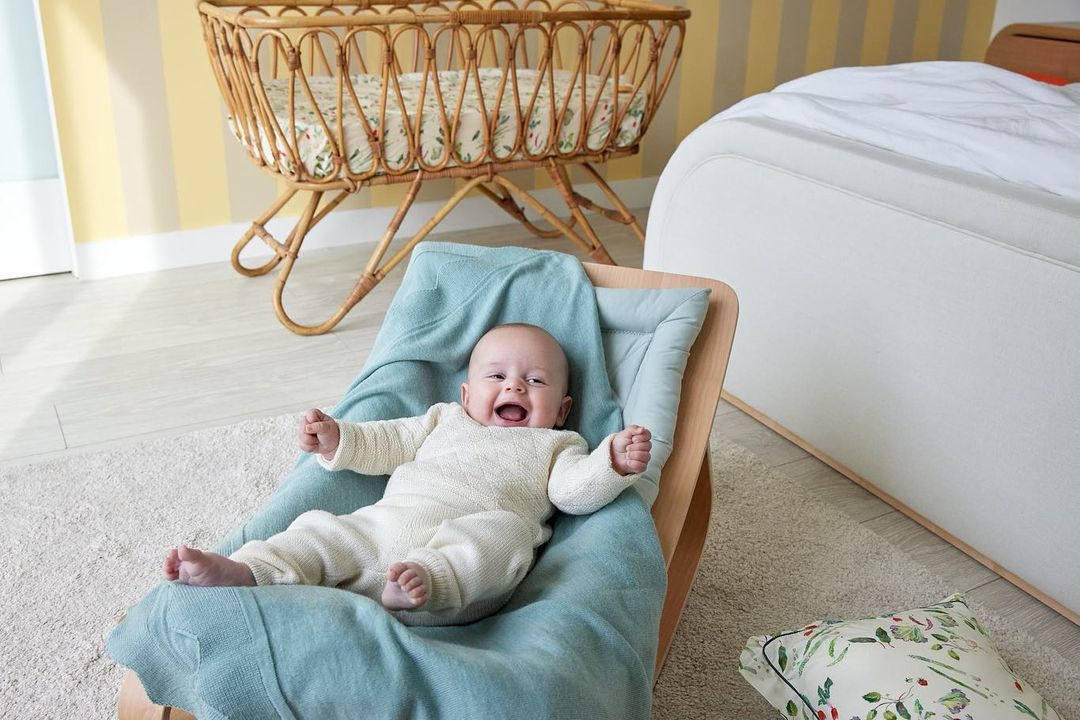 Adorable baby wearing a cream-colored knit onesie, lying in a wooden rocking chair with a light blue blanket, smiling in a well-lit room.