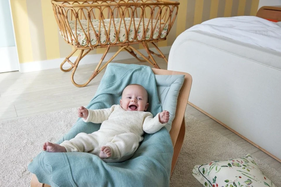 Adorable baby wearing a cream-colored knit onesie, lying in a wooden rocking chair with a light blue blanket, smiling in a well-lit room.