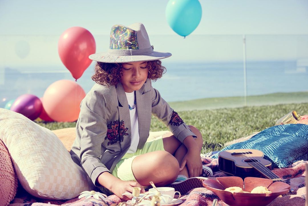 Young girl in a floral blazer and hat sits on a blanket with balloons, tea set, and ukulele, enjoying a picnic on the grass.