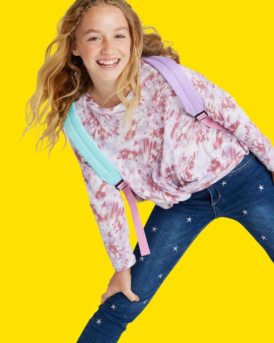Smiling tween girl in tie-dye top and star jeans strikes a playful pose against a solid yellow backdrop, wearing a pastel backpack.