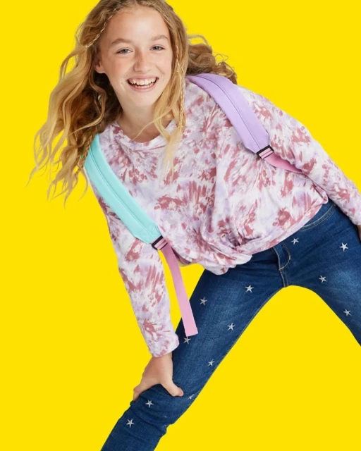 Smiling tween girl in tie-dye top and star jeans strikes a playful pose against a solid yellow backdrop, wearing a pastel backpack.