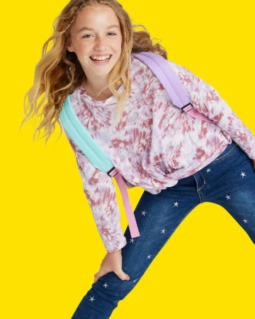 Smiling tween girl in tie-dye top and star jeans strikes a playful pose against a solid yellow backdrop, wearing a pastel backpack.