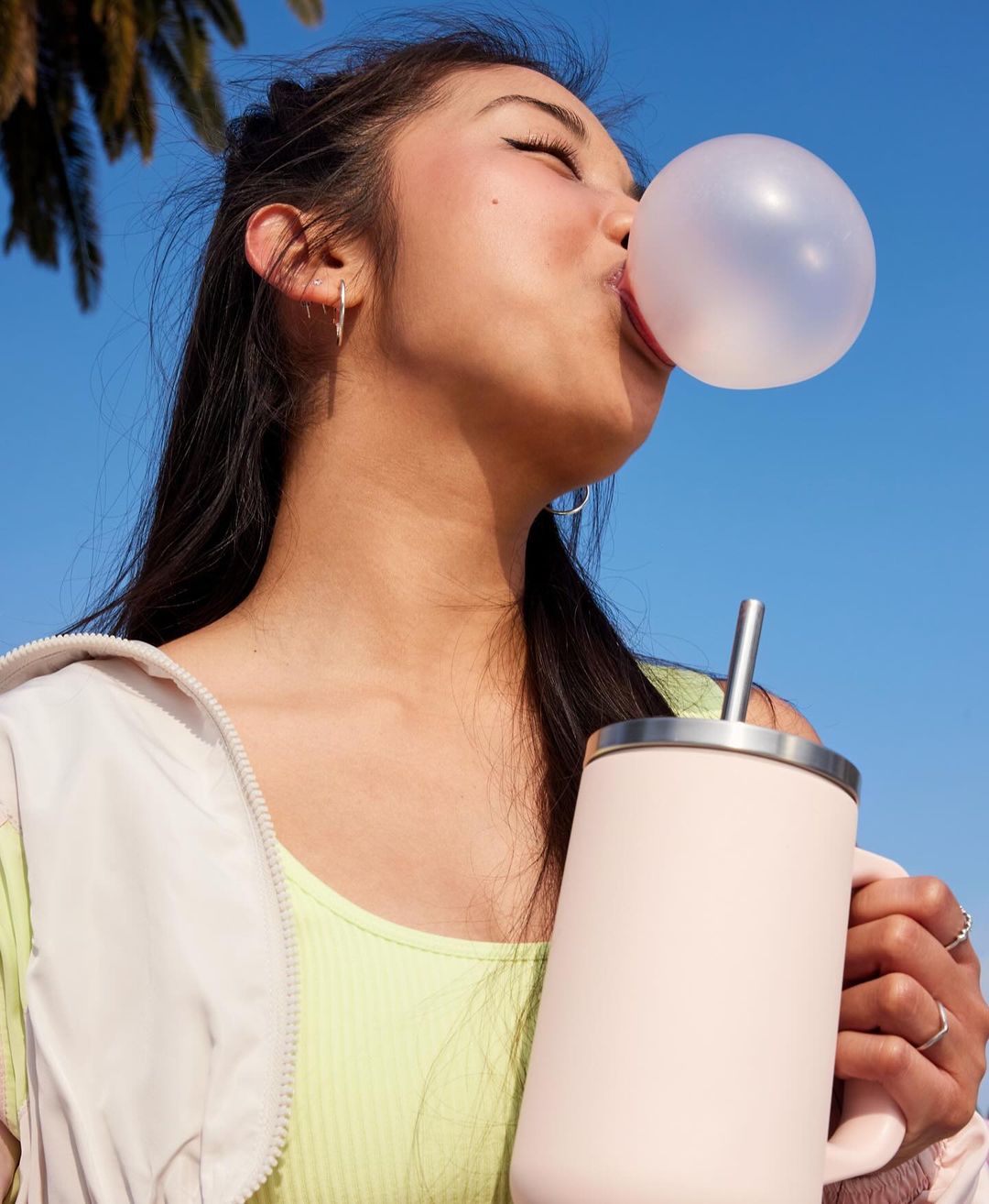 A young adult woman with long dark hair blows a bubble with bubblegum. She wears a light jacket and holds a pink tumbler.