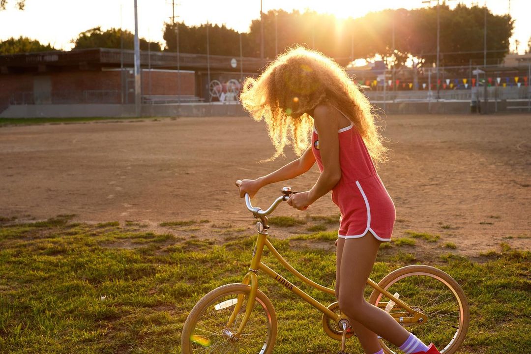 A young woman with curly red hair is riding a vintage yellow bicycle on a grassy field at sunset, wearing a red romper.