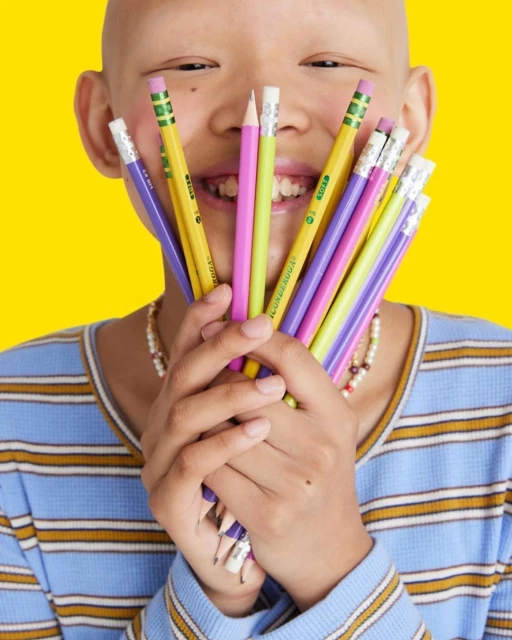 Smiling child with bald head holds colorful pencils against a bright yellow background, wearing a striped blue and tan shirt.