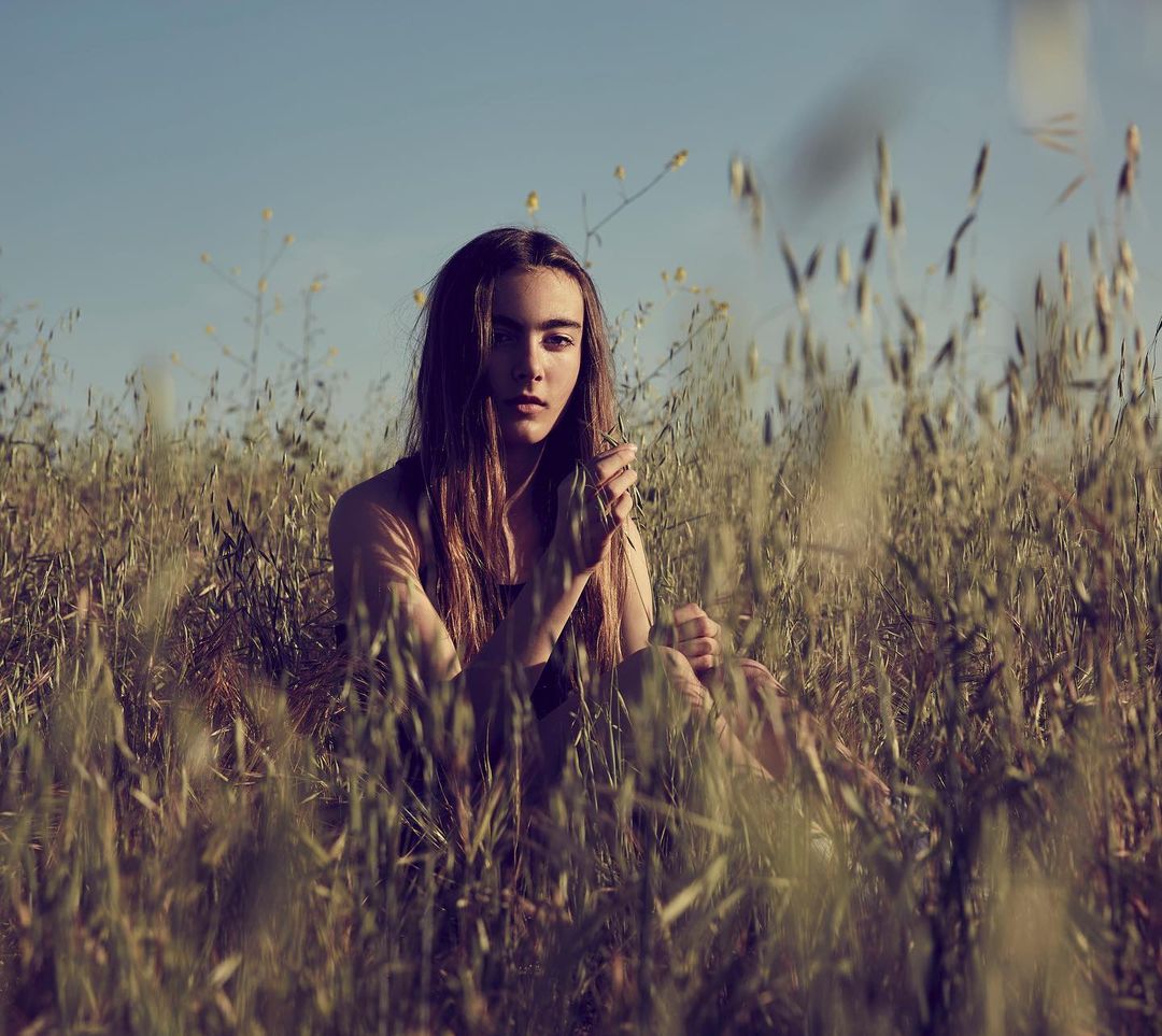 Young woman sits in a field of tall grass under a clear blue sky. She has long brown hair and is holding blades of grass in her hands.