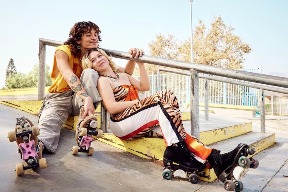 A couple lounges at a skatepark in vintage roller skates and outfits, sitting by metal railings on yellow steps under a bright sky.