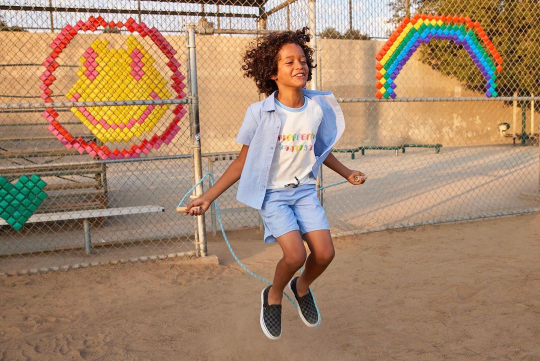 Boy with curly hair jumps rope outdoors on dirt court with colorful shapes on fence in background on a sunny day. 
