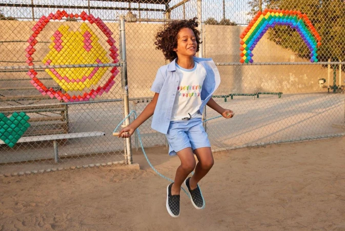 Boy with curly hair jumps rope outdoors on dirt court with colorful shapes on fence in background on a sunny day. 