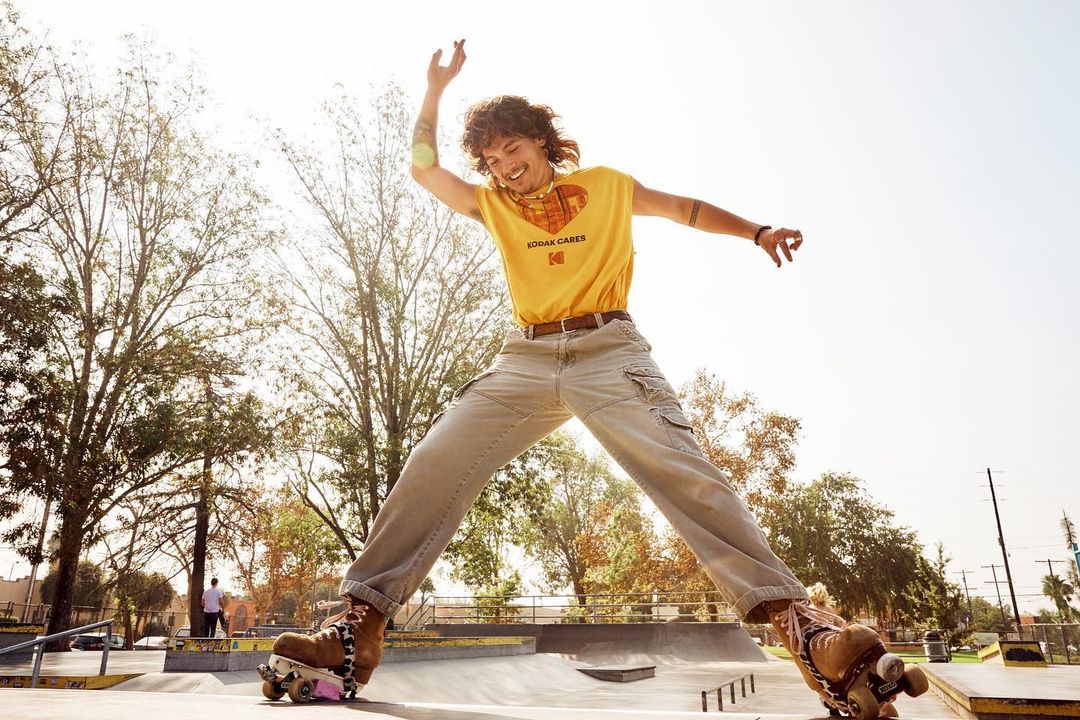 A young adult on roller skates leaps in a sunlit park, wearing a yellow Kodak t-shirt, beige pants, and vintage skates against trees.