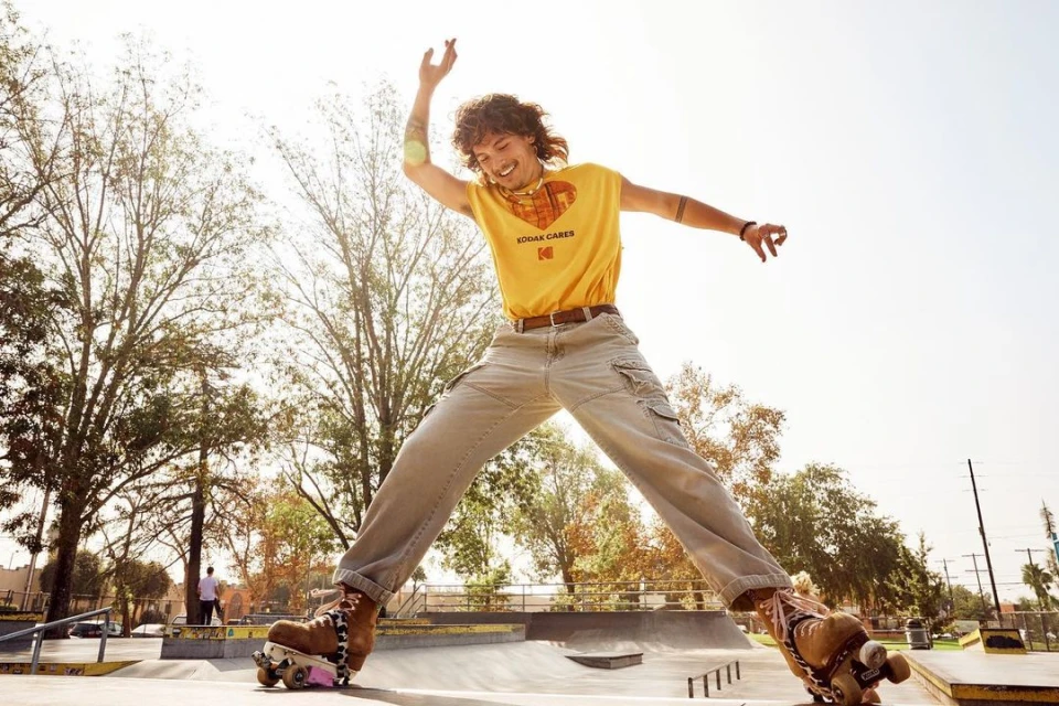 A young adult on roller skates leaps in a sunlit park, wearing a yellow Kodak t-shirt, beige pants, and vintage skates against trees.