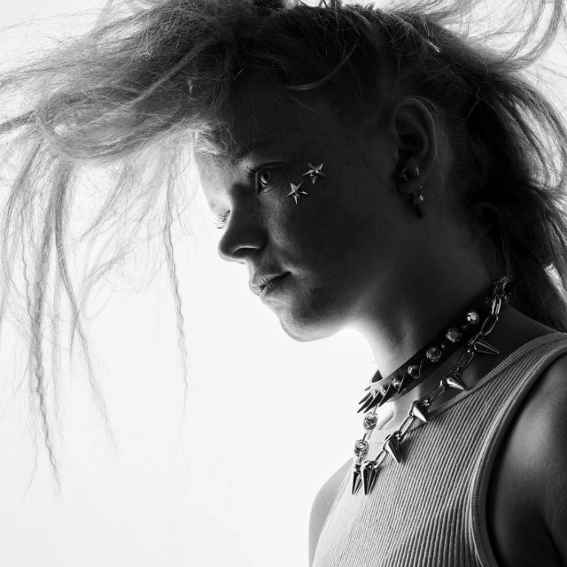 Black and white portrait of a young woman with punk hairstyle, star facepaint, a studded choker, and a sleeveless top against a bright background.