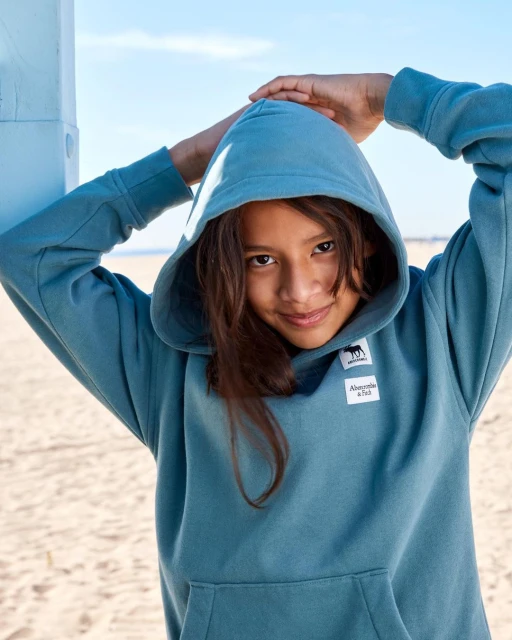 Portrait of a happy tween wearing a blue Abercrombie & Fitch hooded sweatshirt with a logo, posing on a beach with blue sky overhead.