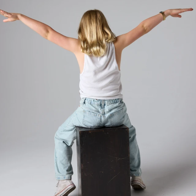 Blond child seated on a cube with arms outstretched against a neutral background in a studio setting, wearing jeans and a tank top.