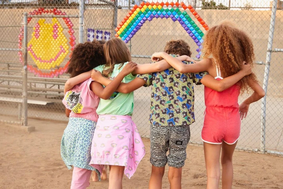 Four children stand arm-in-arm in an outdoor field in front of a chain-link fence decorated with a rainbow and smiley face.