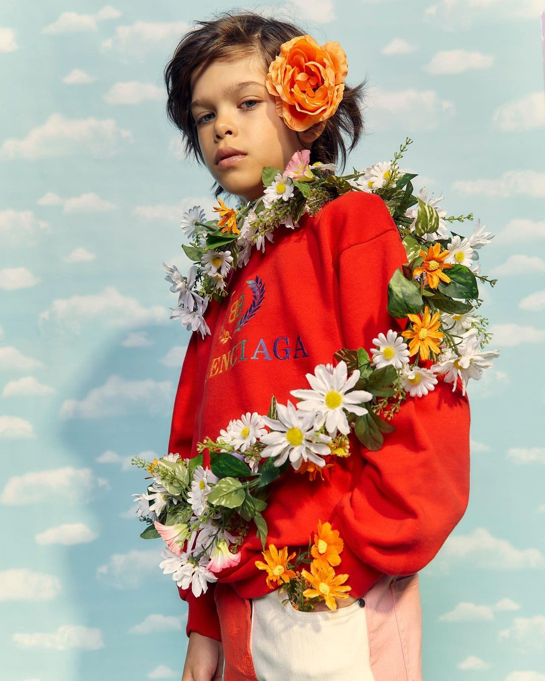 Child model wearing a red Balenciaga sweatshirt surrounded by a daisy floral garland and a orange flower in her hair against a sky background.