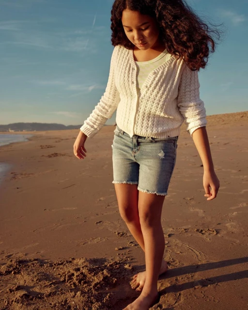 A young barefoot girl in a white cardigan and jean shorts strolls along a sandy beach with the ocean and sky visible in the background.