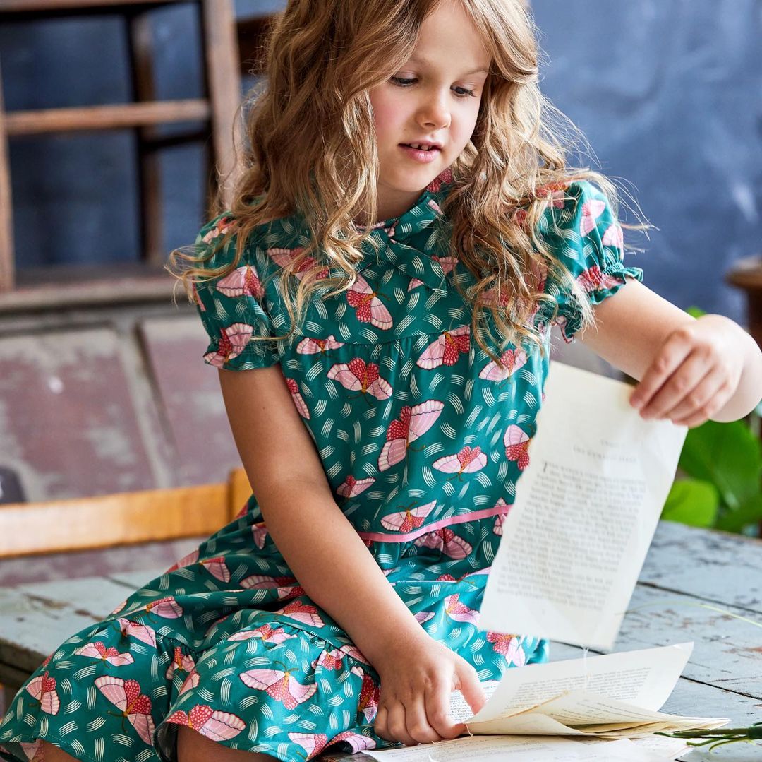 Blonde girl in green patterned dress sits reading papers in an indoor setting with natural light and vintage decor, captured at eye-level.