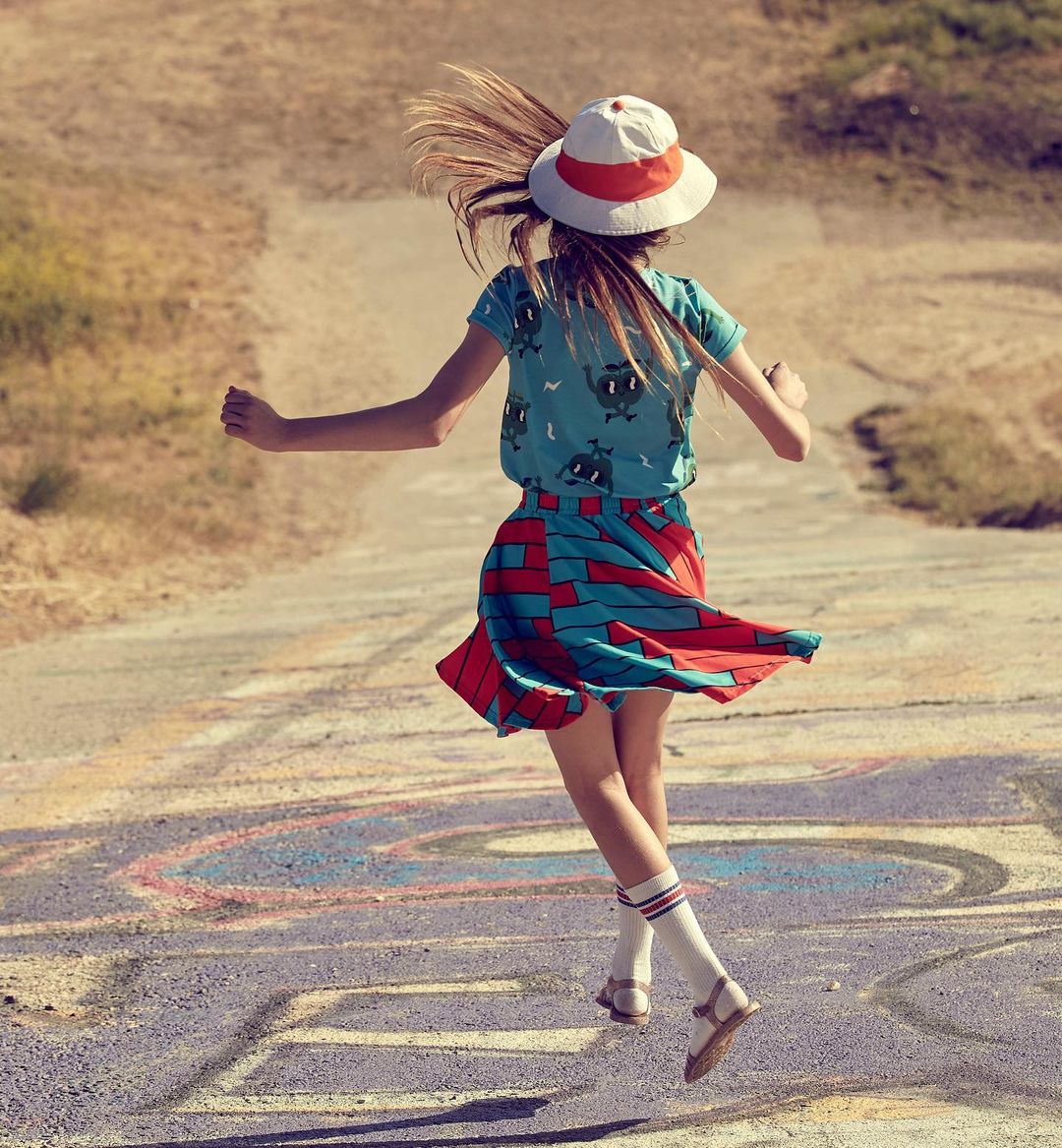 A child in a blue patterned shirt, red skirt, and white hat jumps on an outdoor road with chalk graffiti, expressing energy and fun.