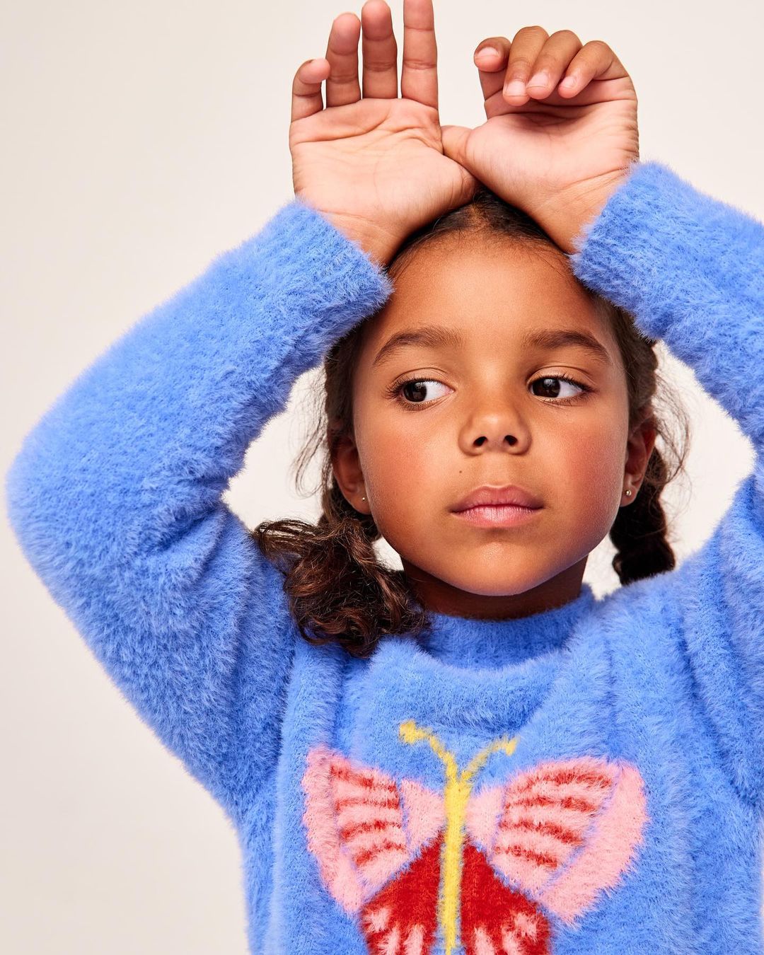 Close-up of a young girl with pigtails wearing a blue sweater with butterfly design, posing indoors with her hands above her head.