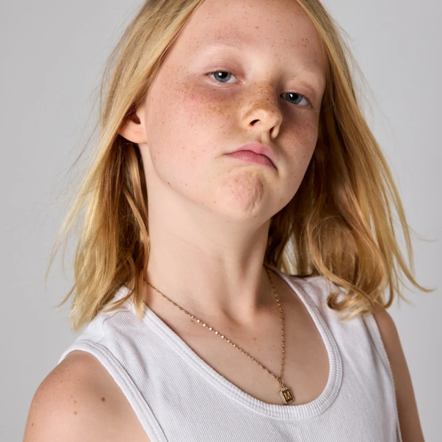 Close-up portrait of a young girl with freckles, blonde hair, and a white tank top against a neutral grey background in a photo studio.
