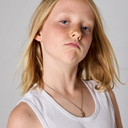 Close-up portrait of a young girl with freckles, blonde hair, and a white tank top against a neutral grey background in a photo studio.