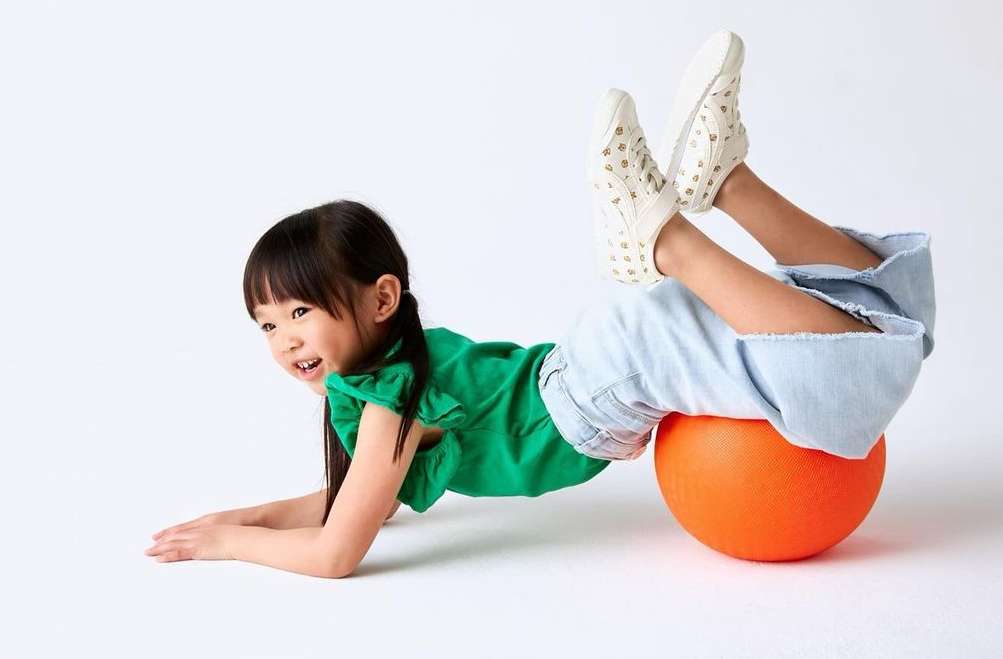 A young Asian girl with dark hair is lying on her stomach on an orange exercise ball. She is smiling and wearing a green top and jeans.