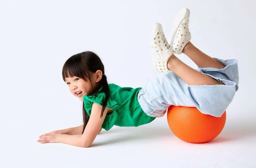 A young Asian girl with dark hair is lying on her stomach on an orange exercise ball. She is smiling and wearing a green top and jeans.