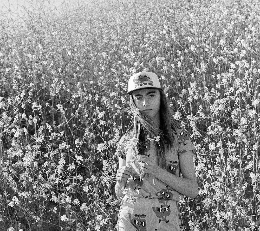 Young woman stands amidst a field of wildflowers in this black-and-white medium shot, wearing a 'California' cap and patterned top.