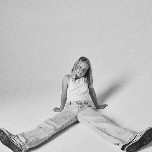 A young girl with blonde hair sits on the floor in a white tank top, wide leg jeans, and black sneakers against a neutral background.