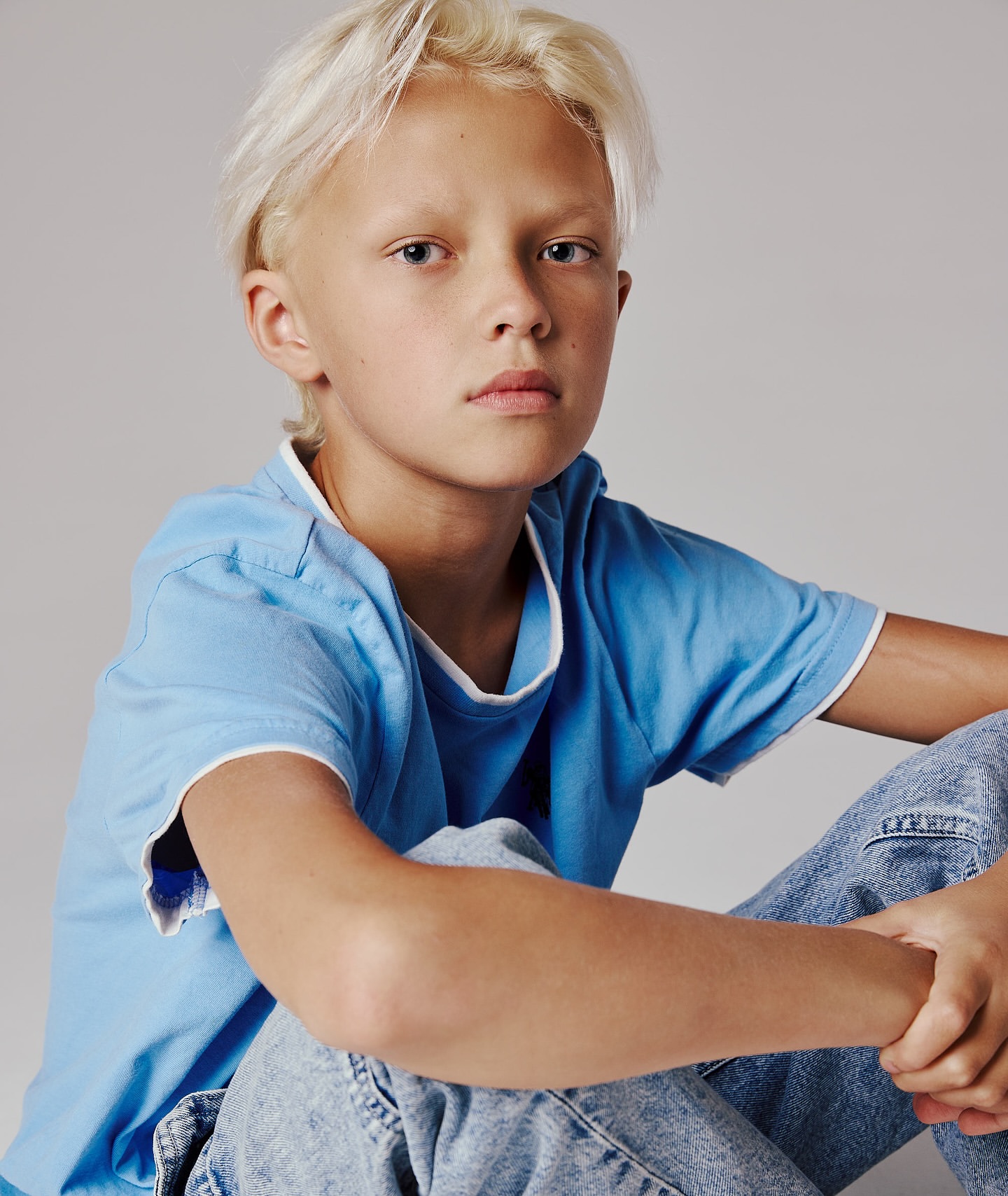 Portrait of a blond child seated wearing a blue shirt and jeans, looking directly at the camera against a neutral grey background.