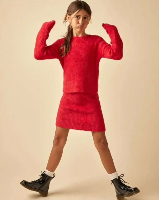 Young girl in a red sweater and skirt makes a funny face while standing indoors, wearing shiny black boots, against a neutral backdrop.