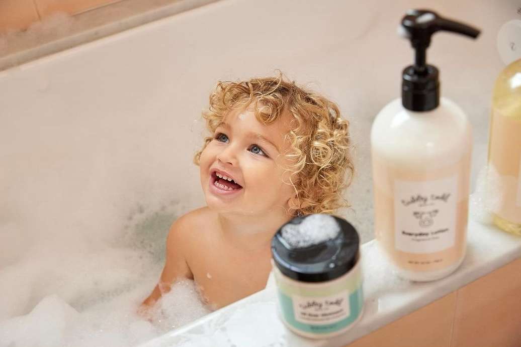 Toddler enjoys a bubbly bath next to personal care product bottles on the side of a tub, looking up with a happy expression.