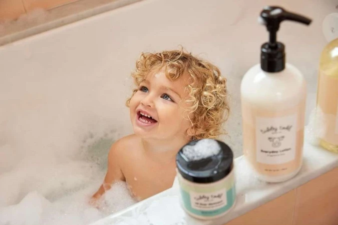 Toddler enjoys a bubbly bath next to personal care product bottles on the side of a tub, looking up with a happy expression.