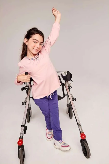 Smiling girl with mobility challenges uses a walker, raising one arm in a bright studio setting, wearing a pink long-sleeve shirt and purple pants.