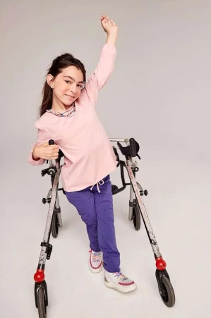 Smiling girl with mobility challenges uses a walker, raising one arm in a bright studio setting, wearing a pink long-sleeve shirt and purple pants.