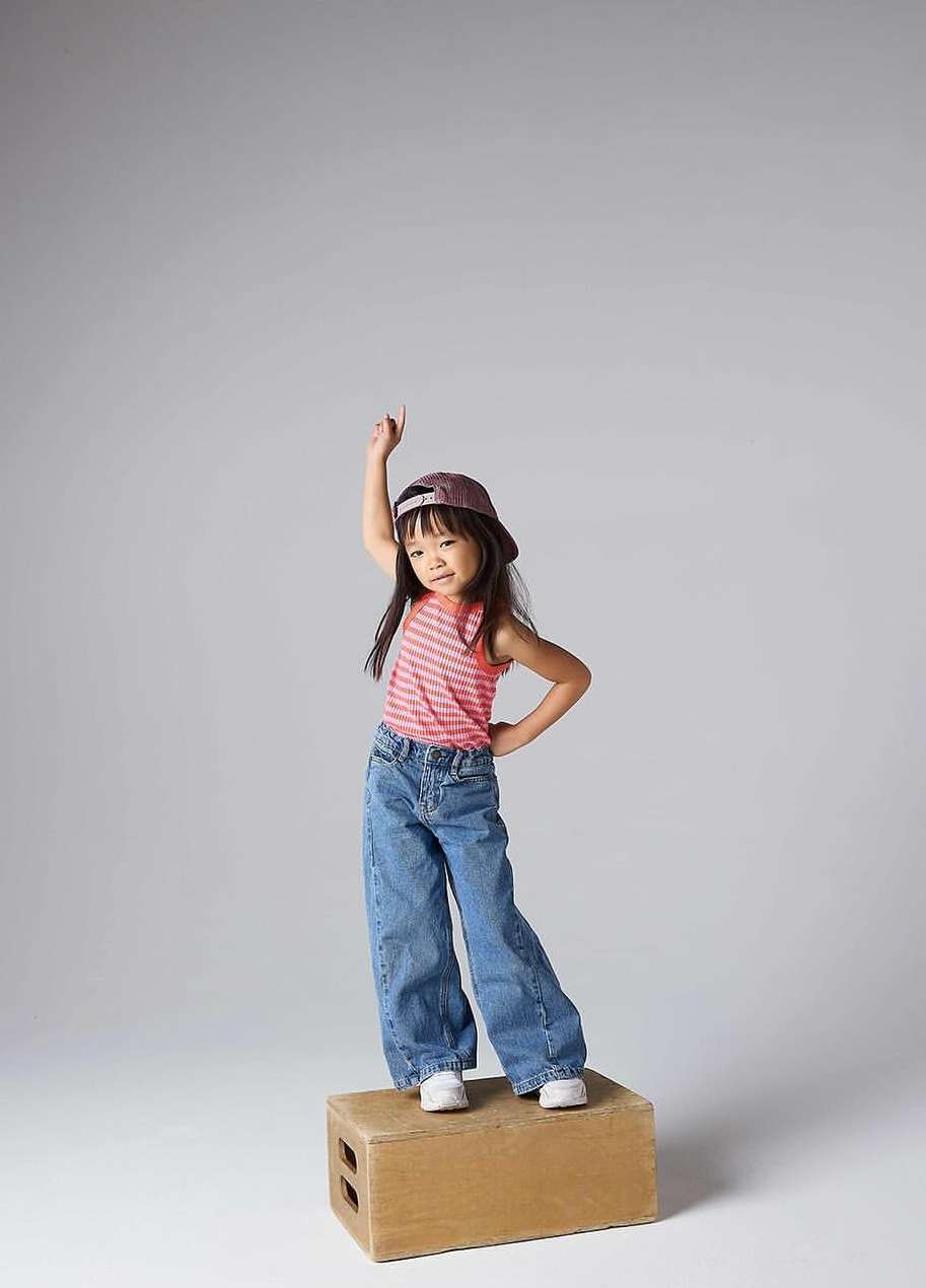 A young girl stands on a wooden block in a photo studio, wearing jeans, a pink top, and a baseball cap, pointing up with one hand.