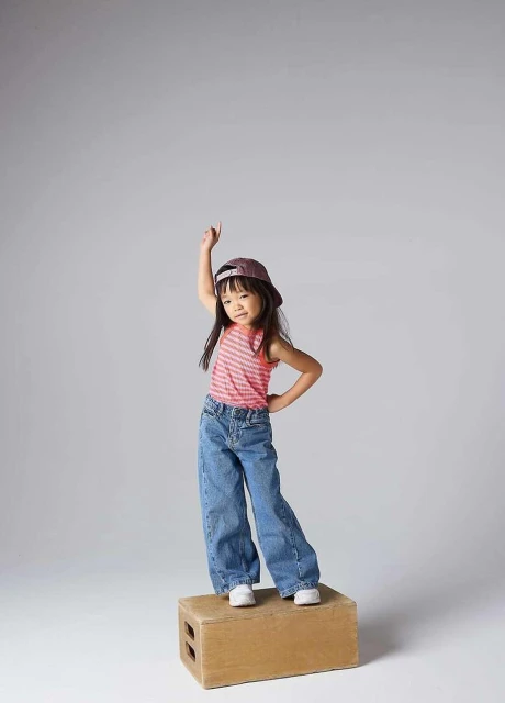 A young girl stands on a wooden block in a photo studio, wearing jeans, a pink top, and a baseball cap, pointing up with one hand.