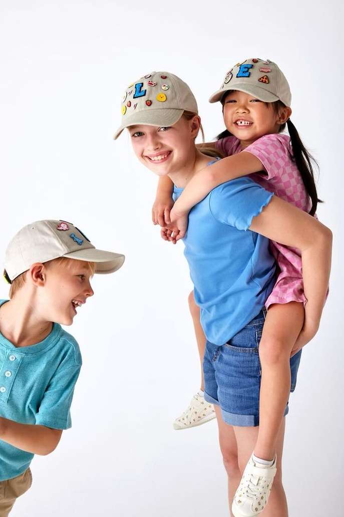 Two smiling girls wearing caps give a piggyback ride in a studio setting, while a laughing boy in cap looks on.