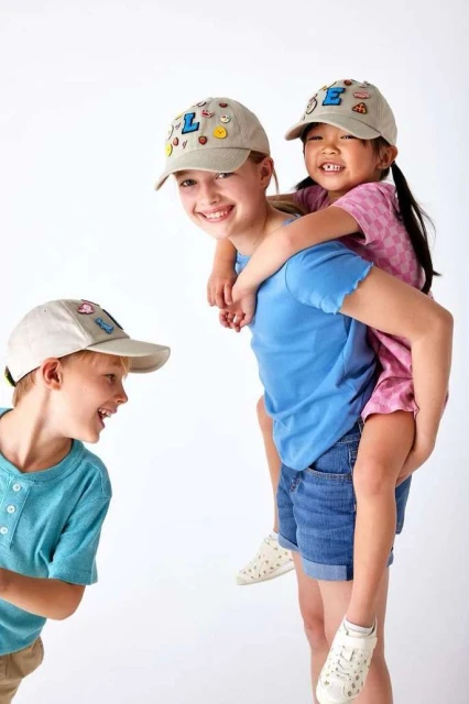 Two smiling girls wearing caps give a piggyback ride in a studio setting, while a laughing boy in cap looks on.