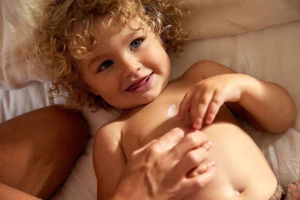 Smiling toddler lying on a white blanket while someone applies cream to their chest, close-up shot, indoor setting with soft lighting.