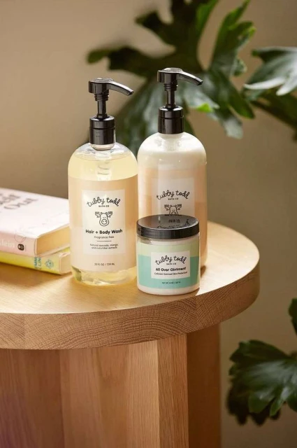 Two Tubby Todd hair and body wash bottles and an All Over Ointment jar on a wooden table, plant leaves in the background.