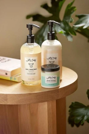 Two Tubby Todd hair and body wash bottles and an All Over Ointment jar on a wooden table, plant leaves in the background.