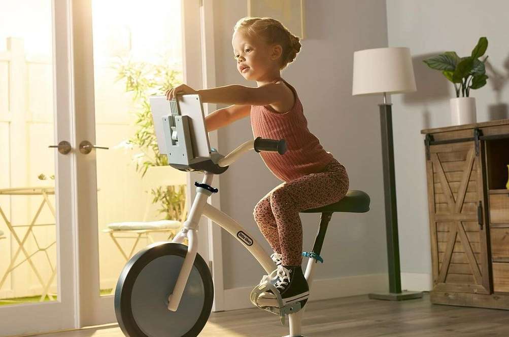 A young child is seated on a stationary exercise bike with a screen attachment, positioned indoors with natural light streaming in.